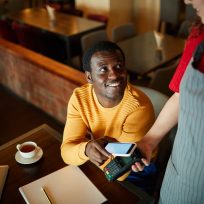 Young smiling man sitting by table and looking at waitress while paying for his order through smartphone