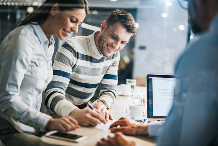 Young happy couple signing a document while being on a meeting with their real estate agent in the office. The view is through glass.