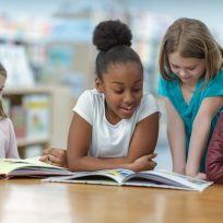 A group of friends are sitting at a table in their classroom. They are reading a book together as they smile and laugh.