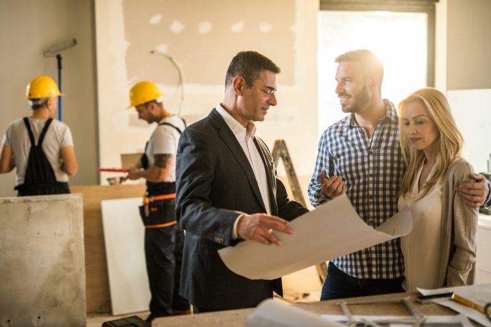 Young happy couple talking to their investor about housing plan inside of an apartment. Workers are in the background.