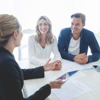 Insurance agent with couple looking through documents. The agent is holding a digital tablet. Couple are casually dressed. They sitting at a table at home and are looking a little happy and smiling.