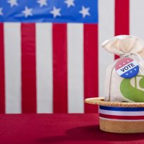 Political Rally Money and the Vote. A skimmer hat sitting on a table with a money bag and a Vote button against a large US flag in the background. Super PAC Money trying to buy vote that will influence the US Elections