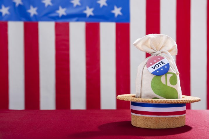 Political Rally Money and the Vote. A skimmer hat sitting on a table with a money bag and a Vote button against a large US flag in the background. Super PAC Money trying to buy vote that will influence the US Elections