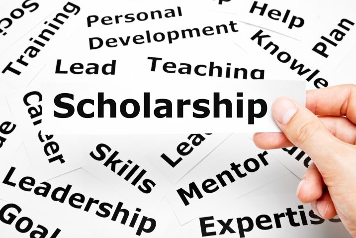 Hand holding a piece of paper with the word Scholarship printed on it above other related words in the background.