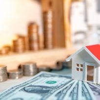 Property or real estate investment concept. Home mortgage loan rate. Saving money for future retirement. Miniature house model with stacked coins and dollar currency banknotes on wooden table.