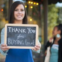 Young business owner holding a "Thank You for Buying Local" sign in front of her store with shopper in the background.