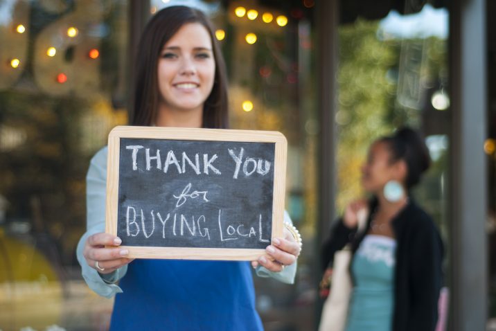 Young business owner holding a "Thank You for Buying Local" sign in front of her store with shopper in the background.