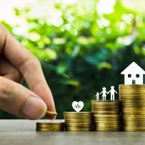 Saving or making money for family life in future concept. A man hand putting coin into rising stack of coins with heart, family and small house model on wood table. Depicts sustainable financial goal.